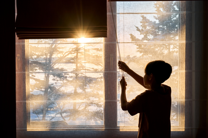 Young boy pulling window blinds up, revealing bright morning sun and snowy pine trees, symbolizing new beginnings and hope